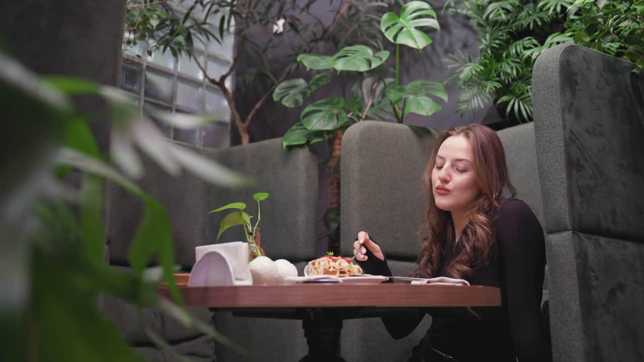 Lady sits in plant-filled cafeteria, peacefully enjoying waffles at table decorated with minimal items and natural accents, creating serene moment of relaxation and indulgence in stylish interior