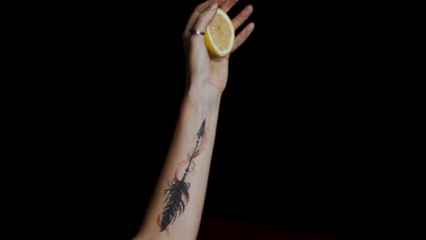 Primer-Plano-De-La-Mano-Femenina-Con-Un-Tatuaje-Exprimiendo-Un-Limón-Sobre-Fondo-Negro