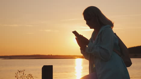 a woman in a hood sits on a fence near a picturesque lake uses a smartphone beautiful sunset