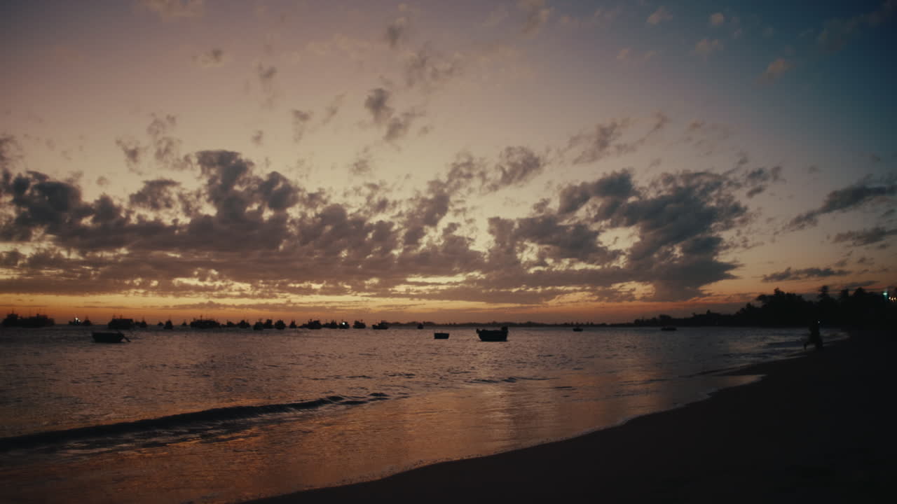 Wide sandy beach with scattered boats and glowing sunset sky over calm ocean water, silhouettes