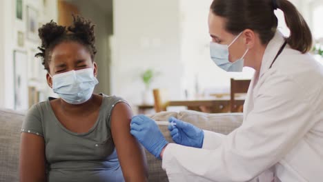 African-american-girl-and-caucasian-female-doctor-wearing-face-masks,-vaccinating