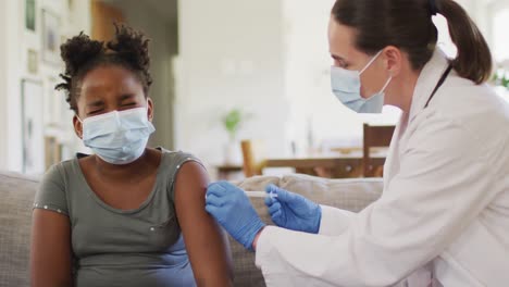 african american girl and caucasian female doctor wearing face masks, vaccinating