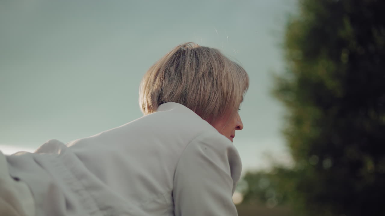 mujer de mediana edad acostada con una expresión enfocada, mirando hacia adelante, la luz natural suave destaca su cara contra un cielo nublado, con árboles en el fondo