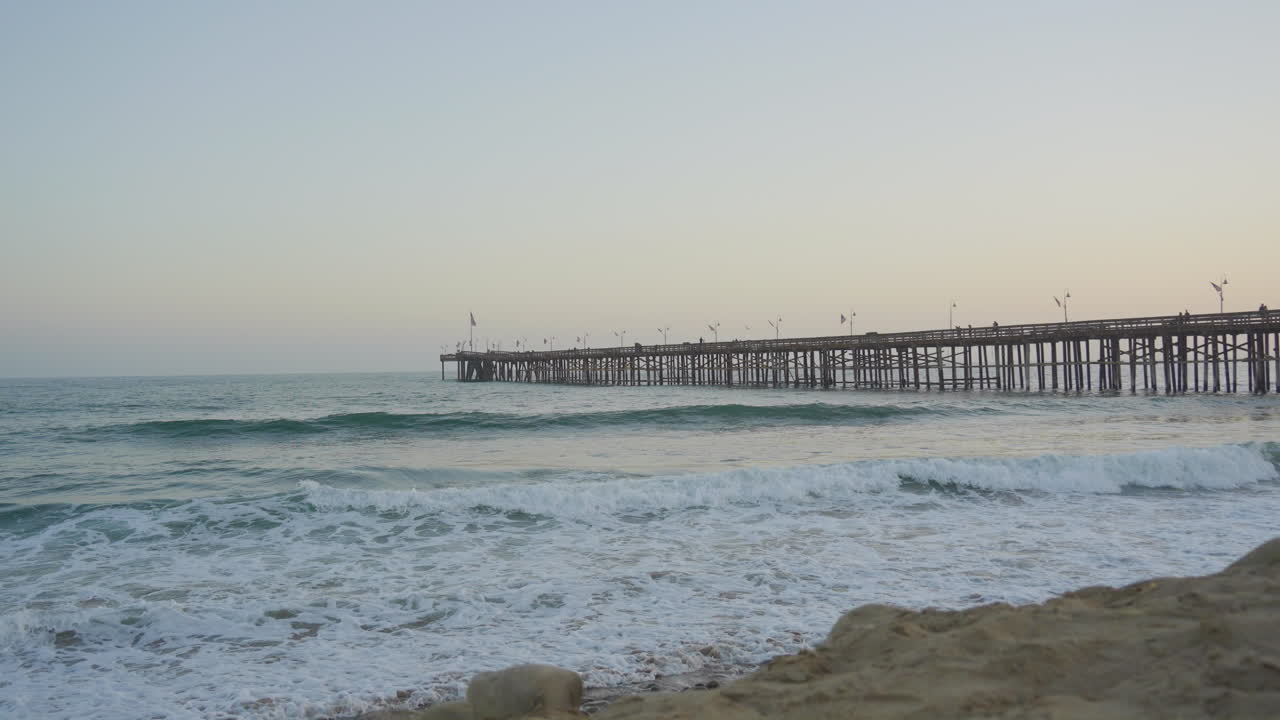 Panoramica al rallentatore di onde che si infrangono dall'Oceano Pacifico sulle coste di Ventura Beach, con il molo e il tramonto sullo sfondo, nella California meridionale