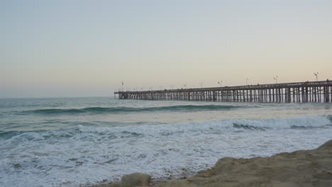 slow motion panning shot of waves rolling through the pacific ocean crashing into the shores of ventura beach with the pier and sunset in the background located in southern california