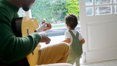 Vista-Lateral-De-Un-Joven-Padre-Negro-Tocando-La-Guitarra-Y-Sentado-En-Una-Silla-En-Una-Cómoda-Casa-4k