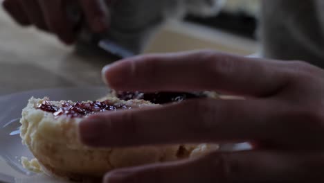 Female-hand-spreads-strawberry-raspberry-jam-from-one-freshly-baked-traditional-scone-to-another-and-uses-knife-for-afternoon-tea-snack