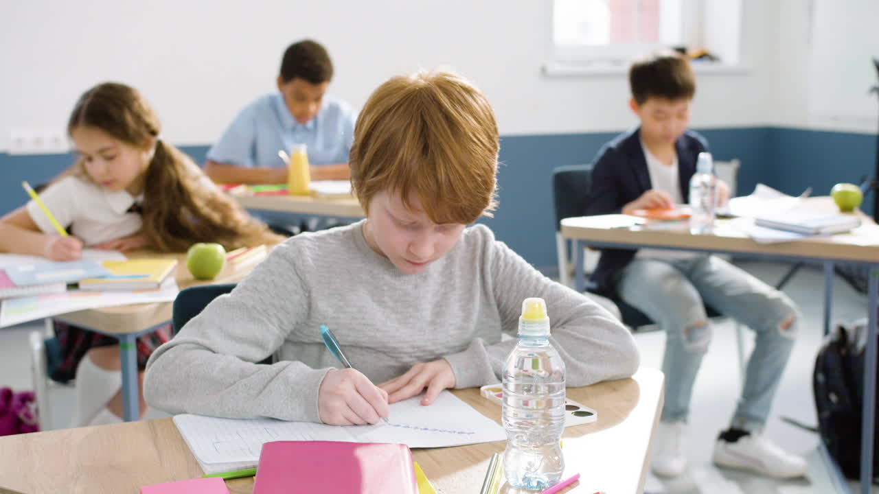 niño pelirrojo sonriente sentado en el escritorio y escribiendo en un cuaderno durante la clase de inglés en la escuela