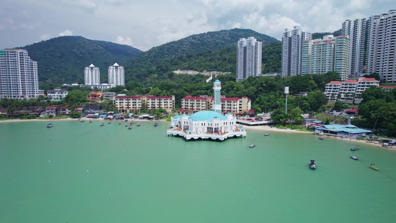 Tanjung Bungah, Penang'daki ikonik mavi kubbeli Yüzen Cami etrafında sinematik havadan dönüş, küçük tekneler, turkuaz-yeşil su, modern yüksek binalar ve ormanla kaplı tepelerle birlikte.