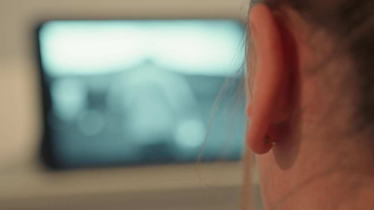 Rear tight view shows person s head and ear angled toward blurred monitor in soft cool light, suggesting concentration, quiet indoor viewing, remote work, study, and calm digital attention