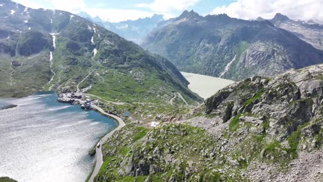 drone pushes towards, stunning blue lake surrounded by green stone mountains, road passes through under cloudy sky at grimselpass, switzerland