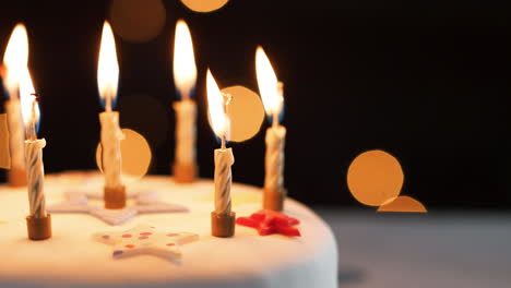 close up of lit candles on a white, decorated birthday cake, which are blown out, bokeh lights in the background