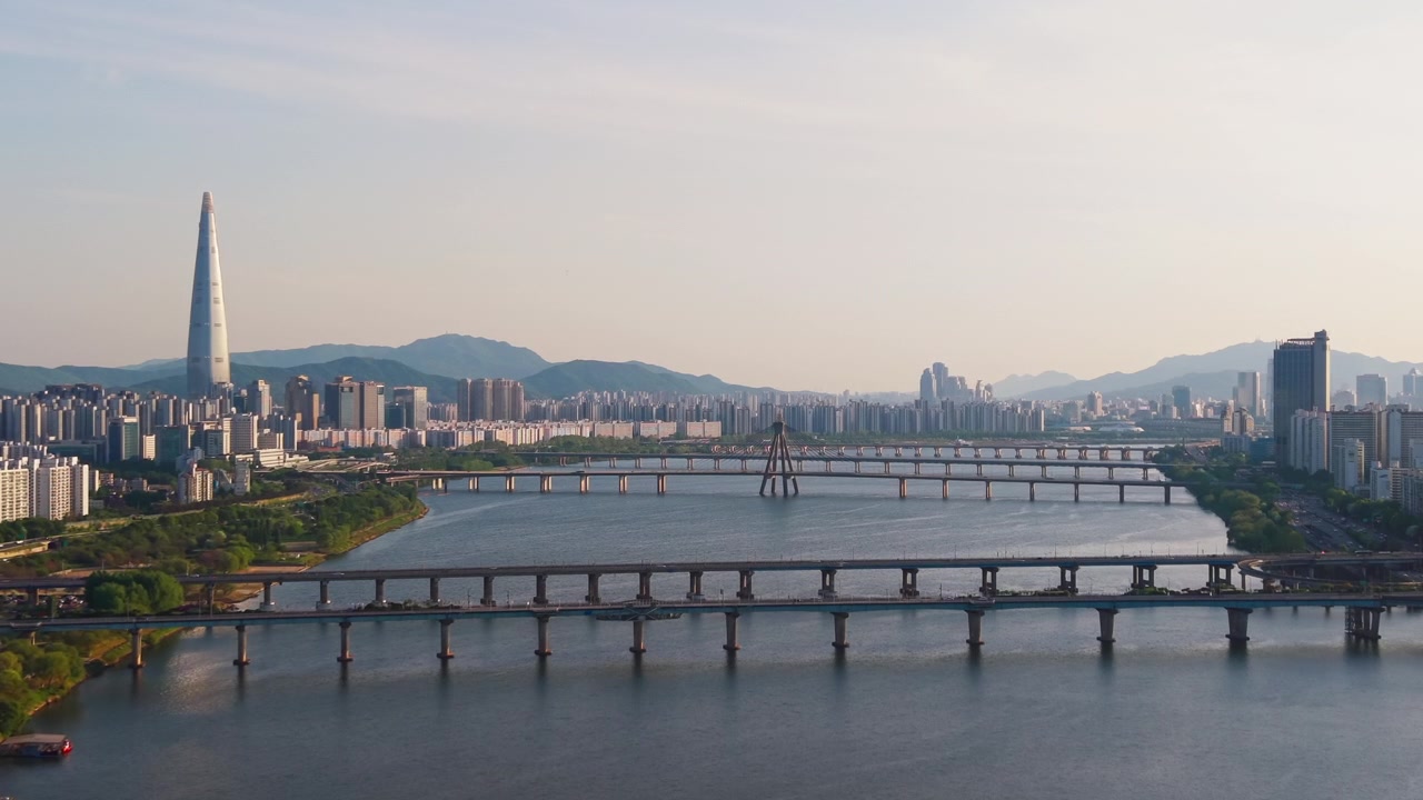 Ripresa aerea lenta in avanti sopra i ponti del fiume Han, vicino al Parco Gwangnaru Hangang, con lo skyline di Seoul e la Lotte World Tower sullo sfondo al tramonto in primavera