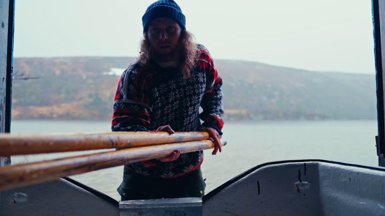 Rowing Boat With A Man Closing The Wooden Door Of A Shed By The Lakeshores. Static Shot