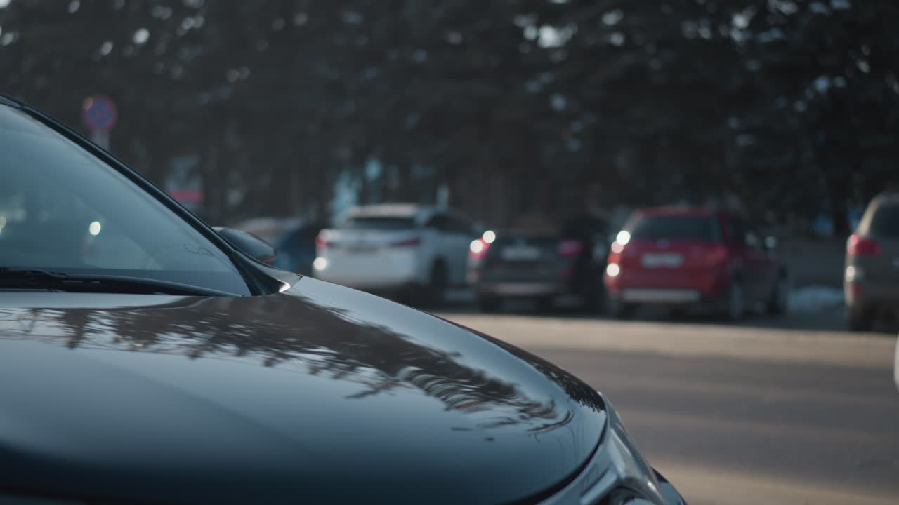 side view of cars parked along both sides of busy urban street with vehicles driving past streetlights and building facades in clear daylight, asphalt pavement, winter season with light snow remnants