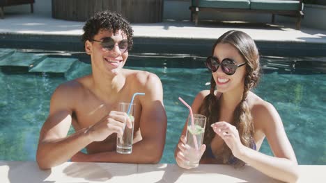 retrato de una feliz pareja biracial con bebidas en la piscina en el jardín en un día soleado