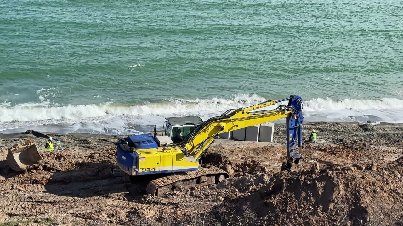 Construction work is taking place near the shore as heavy machinery digs and moves earth along the coastline in a coastal area during daylight hours