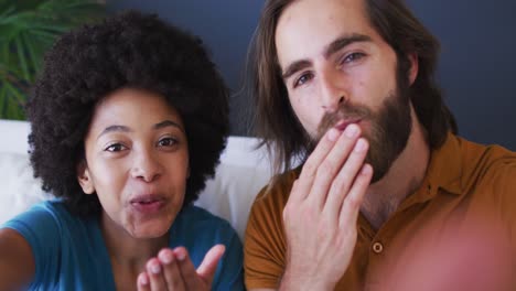 Portrait-of-mixed-race-couple-blowing-kisses-on-videocall-while-sitting-on-the-couch-at-home