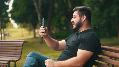 joven apuesto sentado en un banco en el parque riéndose y charlando a través de una videollamada con vista lateral del teléfono celular en cámara lenta