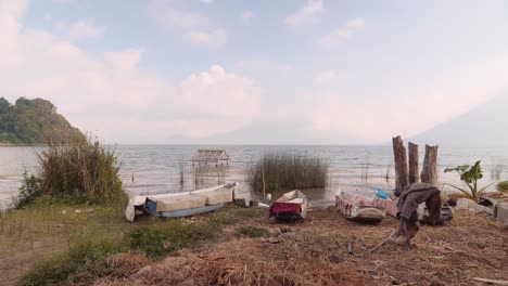 Malerische-Aussicht-Auf-Kleine-Boote-Am-Ufer-Des-Atitlan-Sees-Und-Die-Drei-Mit-Wolken-Bedeckten-Riesen-Im-Hintergrund