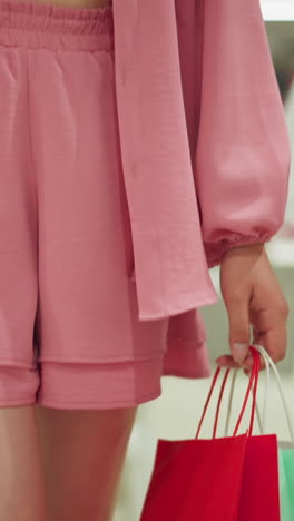 dama blanca con ropa rosa caminando a través de un centro comercial bien iluminado, llevando bolsas de compras verdes y rojas en ambas manos, parece contemplar su próximo paso mientras se mueve a través de la tienda