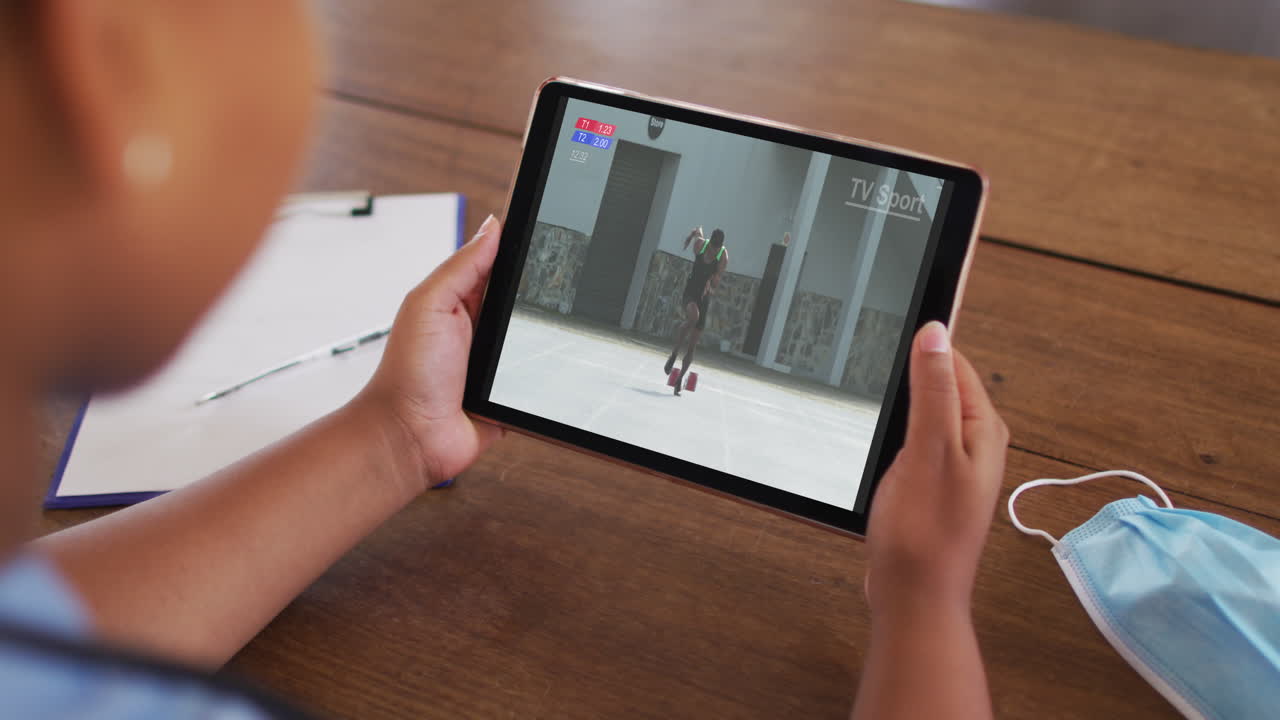 compuesto de una mujer sentada en una mesa con una máscara facial, viendo un evento de atletismo en la tableta