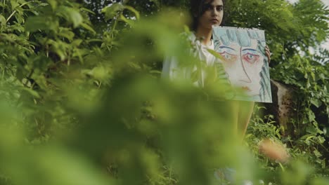 Selective-focus-cinematic-slow-motion-shot-of-a-Young-Asian-happy-girl-sitting-in-nature,-holding-a-painting-while-looking-at-front-of-the-camera