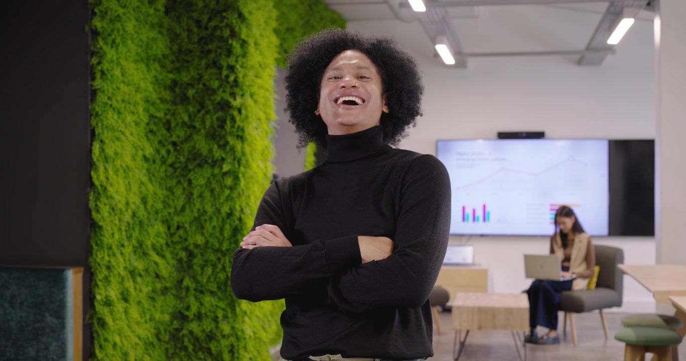 African American team man starting walk along plant wall, posing and smiling while woman on laptop
