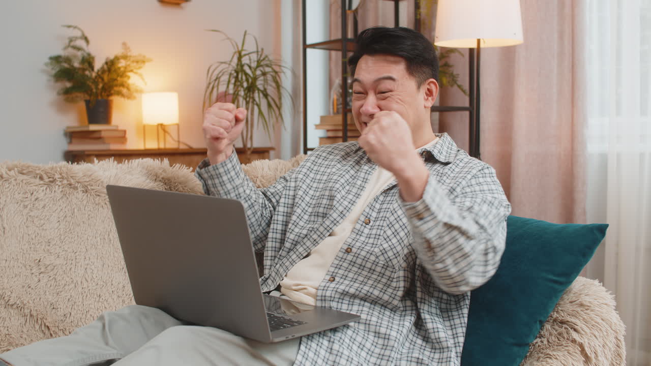 Excited man sitting on sofa using laptop receives good news victory celebrate lottery win at home
