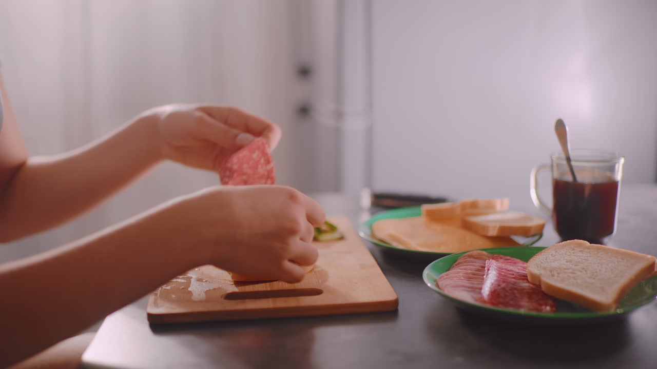 Close up of person preparing sandwich on wooden cutting board in kitchen, arranging slices of bread with cheese and sausage on plates nearby while cup of coffee sits on table