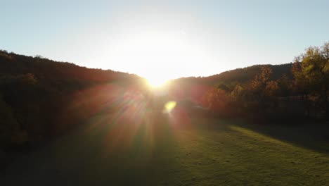 Epische-Drohnenaufnahme-Aus-Der-Luft-Von-Sonnenstrahlen-Auf-Einer-Wiese,-Umgeben-Von-Wald-Bei-Sonnenuntergang