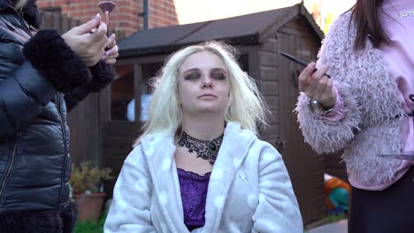 Pale-blonde-girl-having-dark-make-up-applied-for-a-photo-shoot-outside