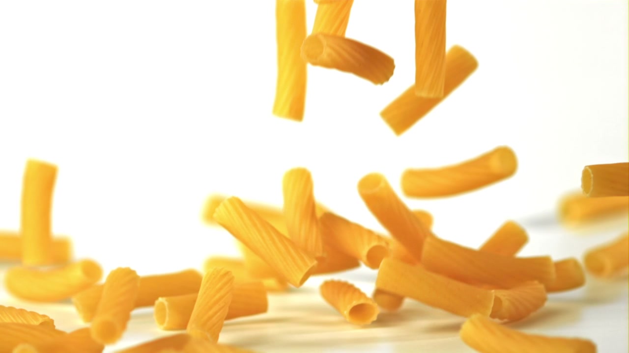 Pezzi di pasta secca dorata che cadono al rallentatore su sfondo bianco.