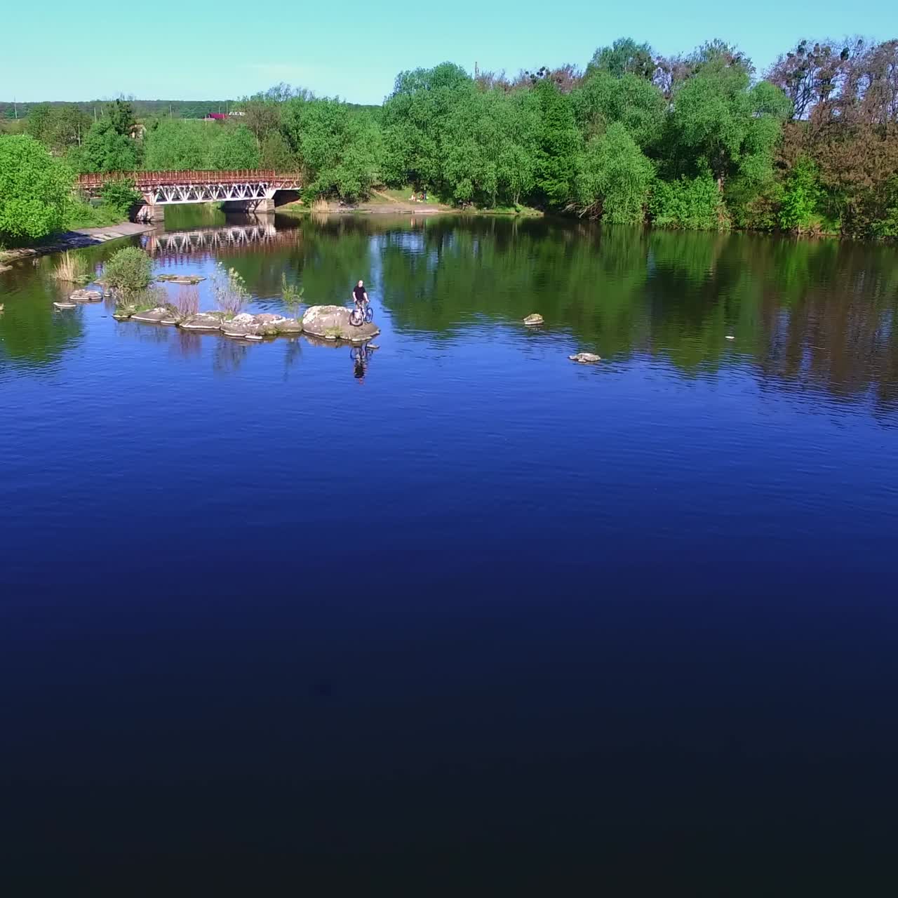 Lovely green wooded bank of the river with metal bridge and house. Man with bicycle standing on the stones peeping from water