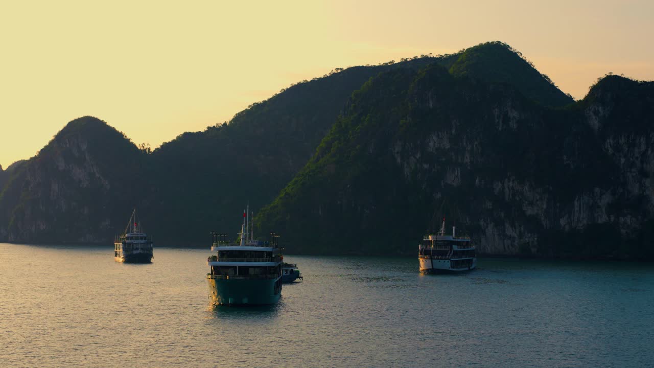grupo de cruceros con siluetas flotando en la bahía de ha long con el cielo naranja del amanecer y las altas montañas en el fondo, viaje a vietnam - pan aéreo revela