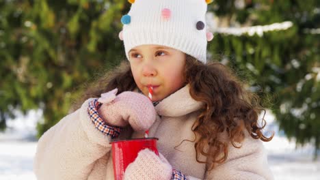 Niña-Bebiendo-Té-Caliente-Durante-Una-Caminata-En-Invierno.-Toma-Aérea-En-ángulo-Bajo-De-Una-Linda-Niña-Con-Gorro,-Parka-Y-Guantes-Bebiendo-Té-Caliente-De-Una-Taza-De-Acero-Inoxidable-Mientras-Camina-Por-El-Bosque-En-Un-Día-Soleado-De-Invierno.