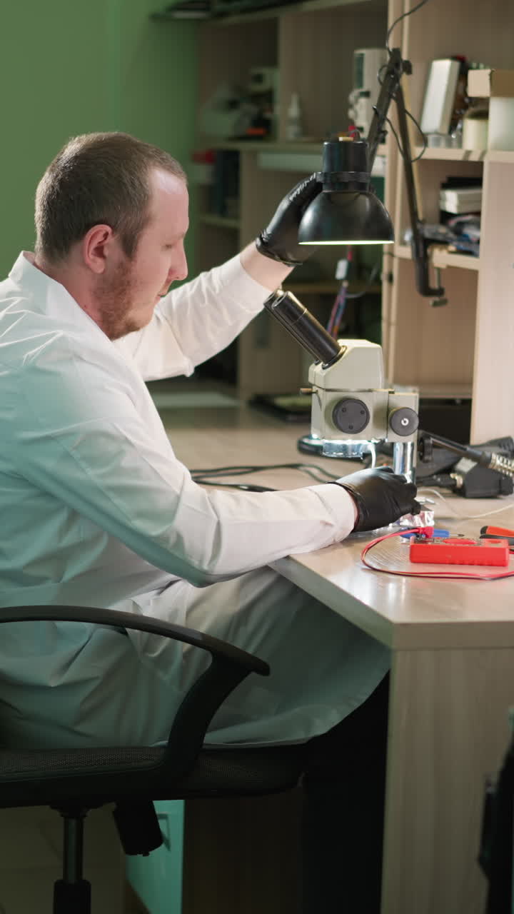 una vista de cerca de un técnico con una bata de laboratorio blanca y guantes de mano negros, ajustando una lámpara de escritorio, el técnico luego continúa trabajando bajo el microscopio, con varias herramientas alrededor