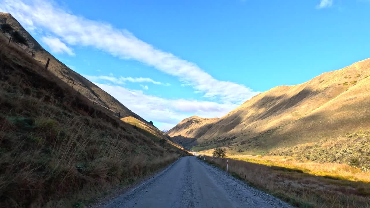 Vehicle drives gravel road between sunlit hills under clear sky, with steady forward camera movement