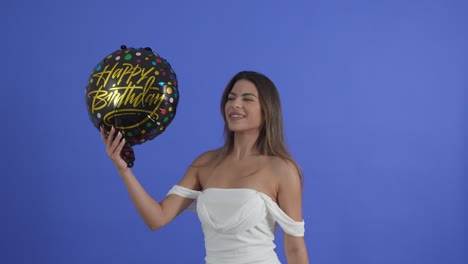 una joven baila con un globo de cumpleaños feliz, aislada en un fondo de estudio azul
