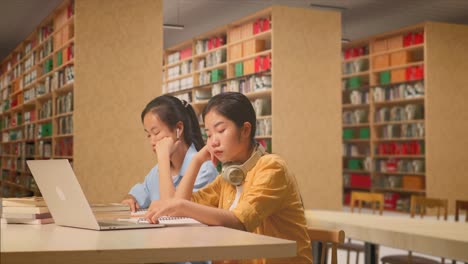 asian woman students with headphones are bored reading the books while sitting on the table with laptop in the library