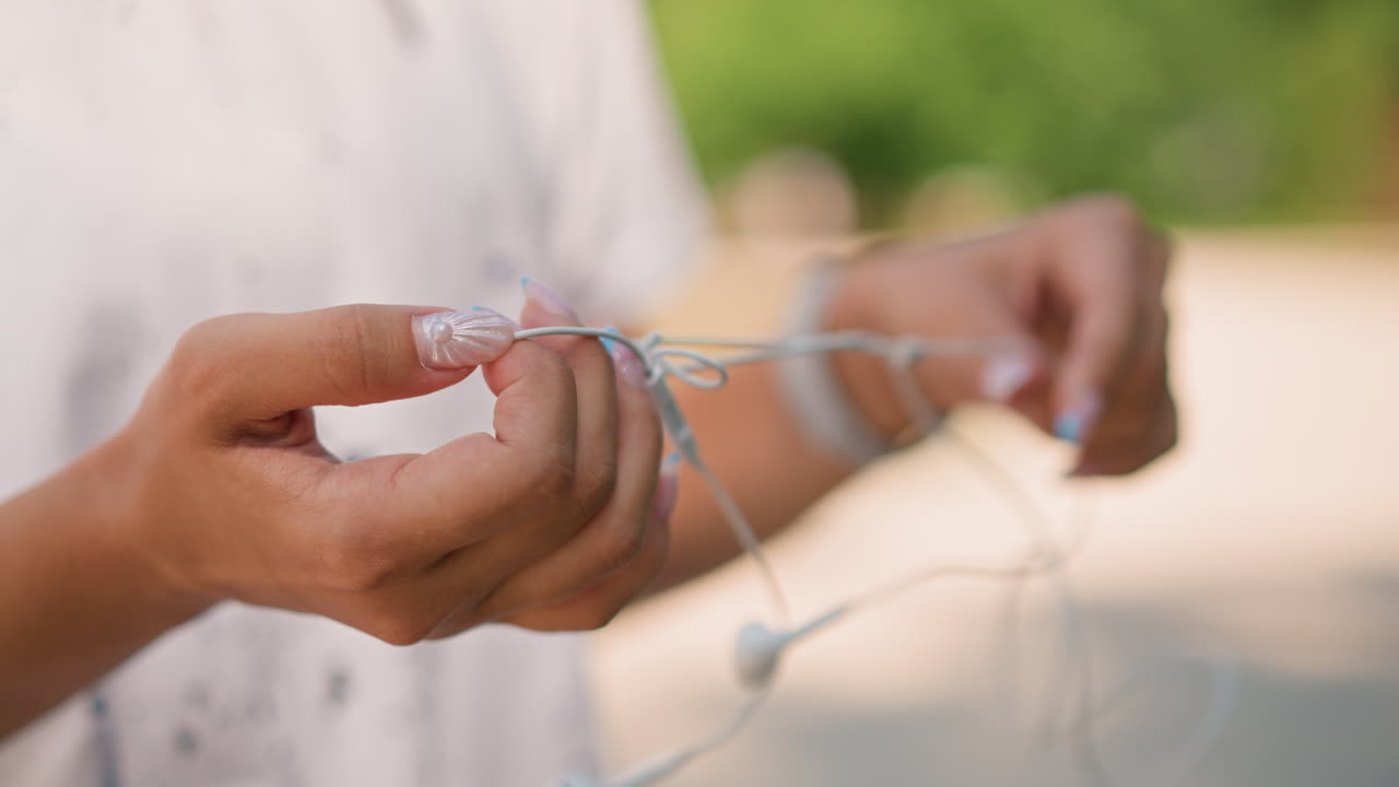 Primer plano de unas manos desenredando unos auriculares blancos enredados; un estudiante de música soluciona el nudo del cable con paciencia; la luz del sol resalta las uñas y la textura; fondo de parque; momento de alivio al desenredar el cable.