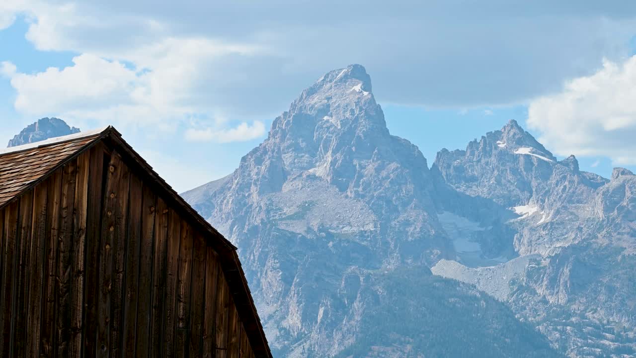 Ein rustikales Holzdach einer Scheune steht im Vordergrund, dahinter erhebt sich dramatisch ein gezackter Berggipfel unter einem teilweise bewölkten blauen Himmel.