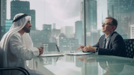 two businessmen with different nationalities sitting in office and signing contract. arab business partner shake hands with investor on financial deal. saudi, emirati, arab businessman concept.