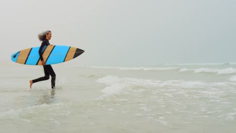 Side-view-of-active-senior-African-American-female-surfer-running-towards-sea-on-the-beach-4k