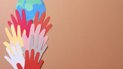 Close-up-of-hands-together-with-globe-made-of-colourful-paper-on-brown-background-with-copy-space