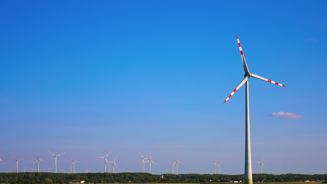 Tall wind turbine rotating in the blue clear sky. Wind farm in the fields at backdrop. Low angle view.
