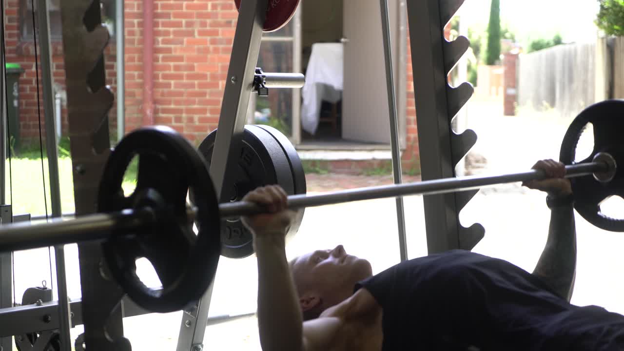 Tattooed man in home gym bench press from the side strong