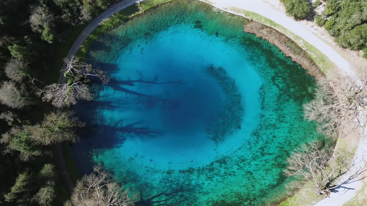 Cinematic drone ascending shot revealing the stunning "Blue Eye" natural spring of Louros. High-angle view of the vibrant turquoise circular lake and crystal clear water. Ideal for nature intros