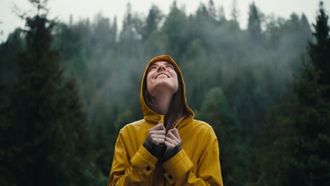 a happy girl in a yellow jacket smiles and waits for the coming rain against the backdrop of a green coniferous forest in the mountains