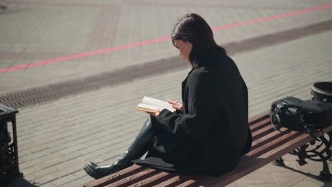 woman in black coat sitting on bench outdoors reading a book, flipping to a new page, her bag rests on the bench beside her, with city streets and sunlight in the background